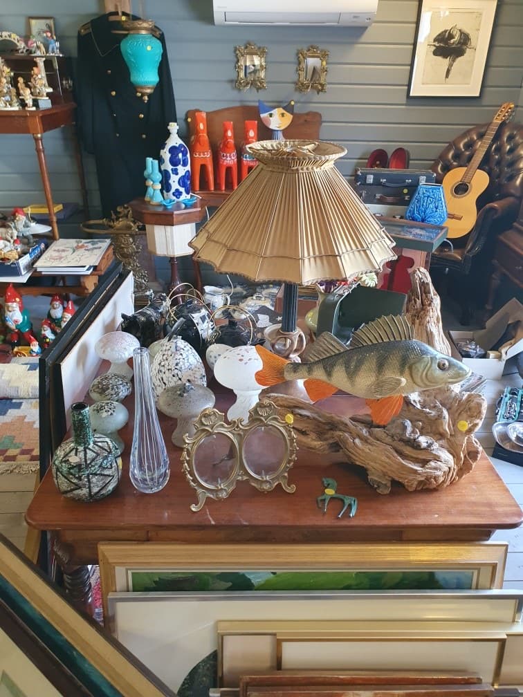 Bordslampa med tygskärm och en träskulptur i form av en fisk, med gitarr och tomtefigurer i bakgrunden